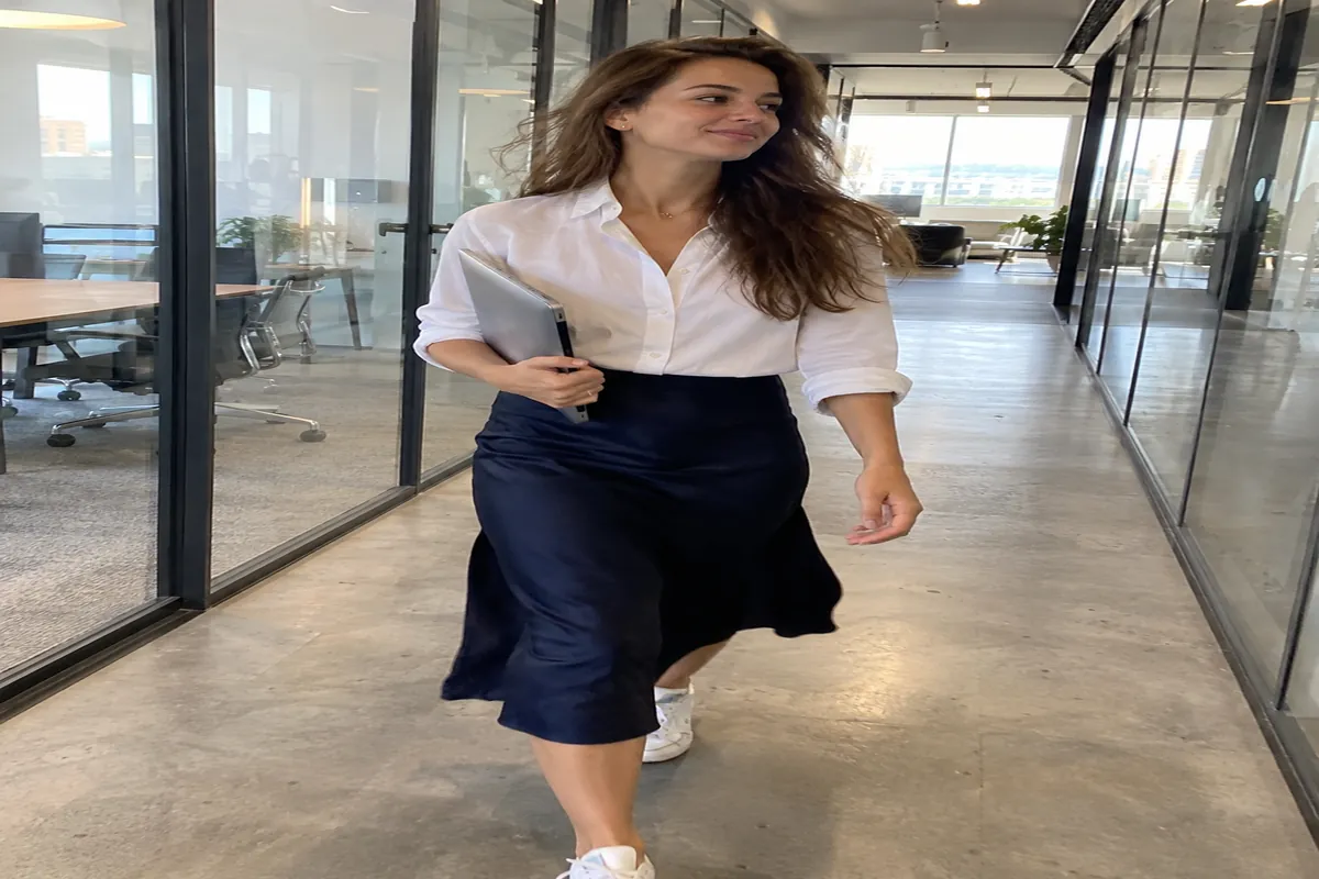 A woman in a navy midi skirt and white shirt walks through a glass-walled office corridor